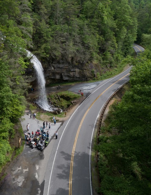 A waterfall cascades down a rocky cliff beside a winding road, with a group of people gathered below.