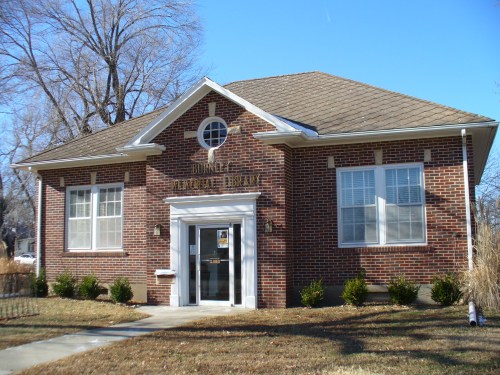A brick building with a gabled roof, labeled "Burnet Public Library," surrounded by grass and small shrubs.