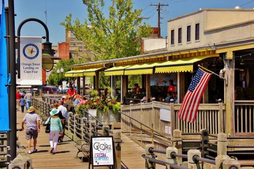A bustling boardwalk scene with people walking, shops, and an American flag, set against a sunny backdrop.