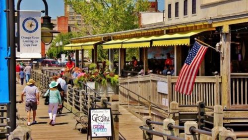 A bustling boardwalk with people walking, shops, and an American flag near the water. Bright sunny day.