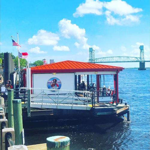 A riverside restaurant with a red roof, flags flying, and a bridge in the background under a bright blue sky.