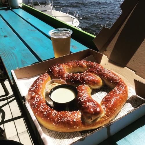 A large, soft pretzel with salt and dipping sauce, alongside a beer, on a table by the water.