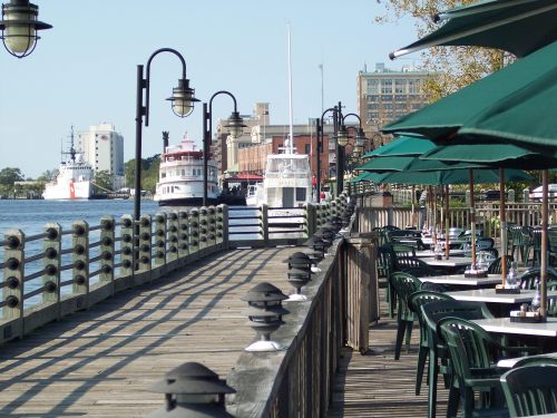 A waterfront boardwalk with tables and chairs, boats docked nearby, and buildings in the background under a clear sky.