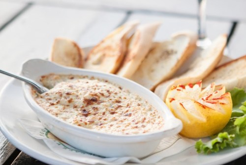 A creamy baked dish served with toasted bread and a grilled lemon slice on a white plate.