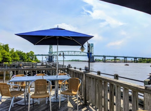 A riverside deck with a blue umbrella, tables, and chairs, overlooking a bridge and calm water under a clear sky.