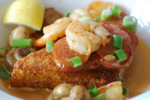 A close-up of a dish featuring fried fish topped with shrimp, sausage, mushrooms, and green onions, garnished with lemon.