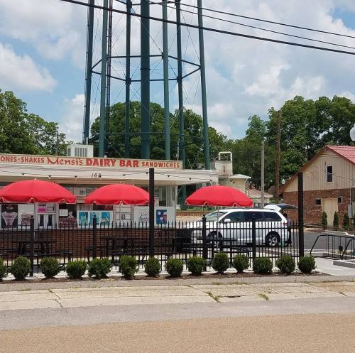 A dairy bar with red umbrellas outside, near a water tower and green trees, on a sunny day.