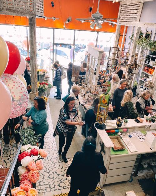 A busy flower shop filled with customers, colorful balloons, and vibrant floral arrangements. Bright orange walls enhance the scene.