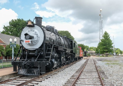 A vintage steam locomotive parked on a railway track, surrounded by greenery and under a partly cloudy sky.