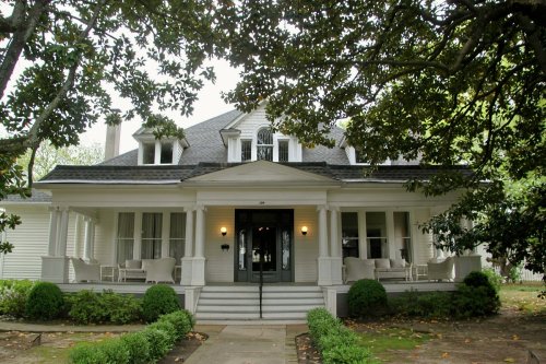 A charming white house with a large porch, surrounded by greenery and shaded by trees.
