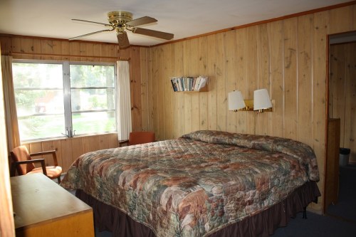 Cozy bedroom with wooden walls, a large bed with a patterned quilt, a fan, and a window letting in natural light.