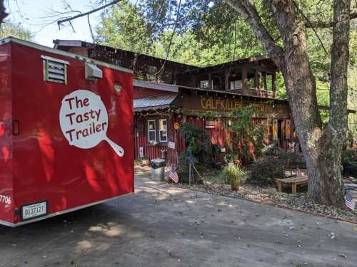 A red food trailer named "The Tasty Trailer" parked near a rustic building surrounded by trees and decorations.