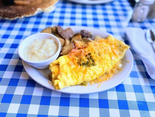 A plate of fluffy omelette with cheese, served with seasoned potatoes and a side of grits on a blue checkered tablecloth.