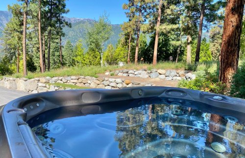 A hot tub with clear water overlooks a scenic view of trees and mountains under a blue sky.