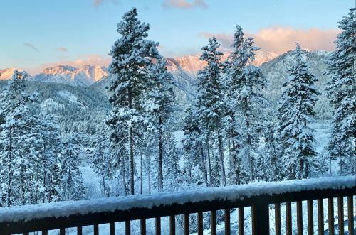 Snow-covered mountains and trees under a clear blue sky, viewed from a wooden balcony.