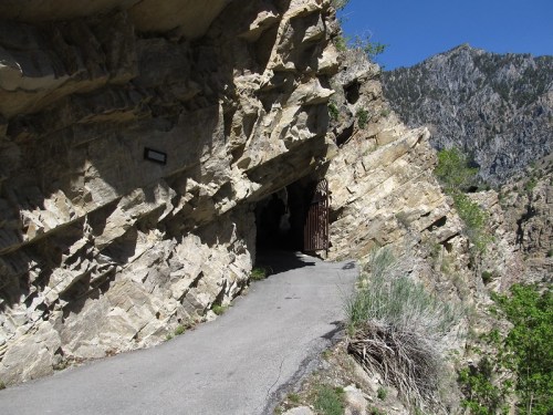 A narrow path leads through a rocky cliff, with mountains in the background and clear blue skies above.