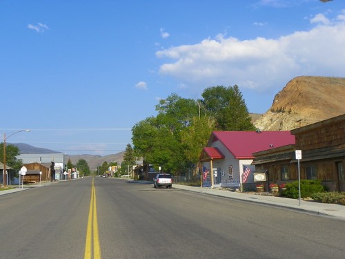 A quiet street in a small town, lined with buildings and trees, under a blue sky with scattered clouds.