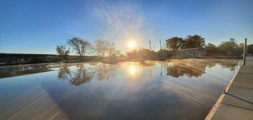 A serene sunrise over a calm lake, reflecting trees and buildings, with mist rising from the water's surface.