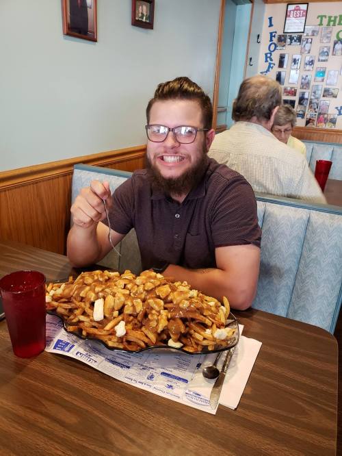 A smiling man with glasses sits at a table with a large plate of poutine, surrounded by diners in a cozy restaurant.