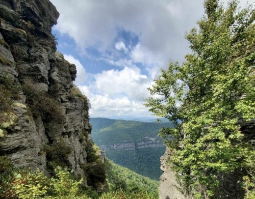 A scenic view of mountains and valleys, framed by rocky cliffs and lush greenery under a partly cloudy sky.