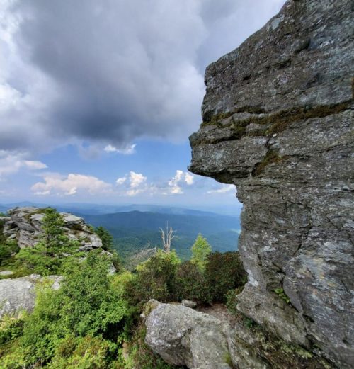 A rocky outcrop overlooks a lush green valley under a cloudy sky, showcasing a scenic mountain landscape.