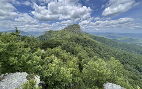 A panoramic view of lush green mountains under a partly cloudy sky, featuring a prominent rocky peak in the distance.