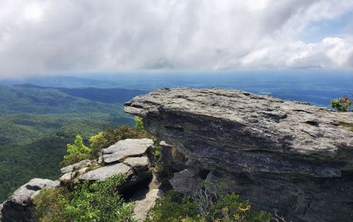 A rocky outcrop overlooks a lush green valley under a cloudy sky, with distant mountains visible in the background.