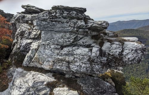 A rugged rock formation with layered textures, surrounded by a forested landscape and distant mountains.