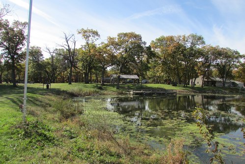 A serene park scene featuring a pond, green grass, trees with autumn foliage, and a gazebo in the background.