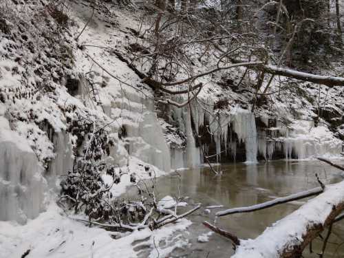 A snowy landscape featuring icy cliffs and frozen waterfalls beside a partially frozen stream.