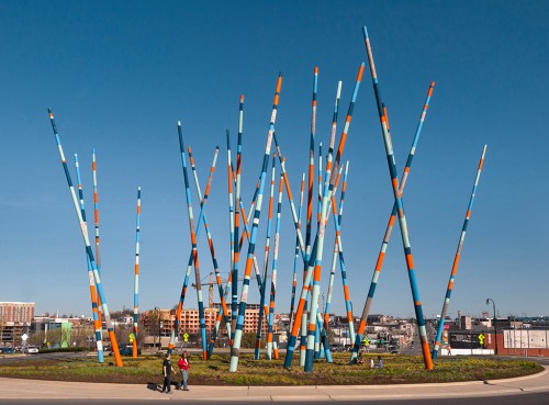 Colorful, tall poles in various heights stand in a grassy area against a clear blue sky, with people walking nearby.