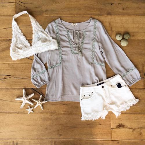 A light gray blouse, white shorts, and a white bralette laid out on a wooden surface with seashells and starfish.