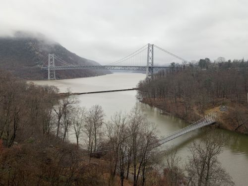 A view of a bridge spanning a river, surrounded by foggy mountains and bare trees in a tranquil landscape.