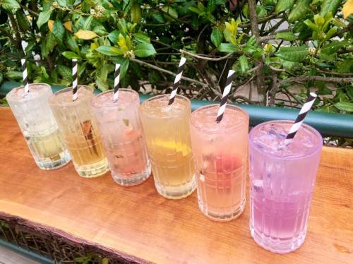Six colorful drinks in tall glasses with black and white striped straws, lined up on a wooden table against a green backdrop.