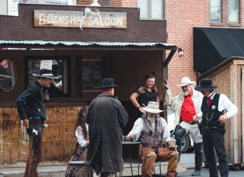 A group of people in Western attire gather outside the Buckshot Saloon, engaging in conversation and enjoying drinks.
