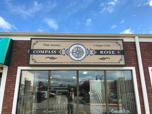 Sign for "Compass Rose" featuring fine jewelry and unique gifts, with a compass design and storefront reflection.