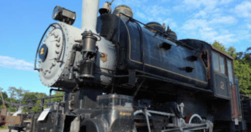 A vintage steam locomotive with a black exterior and brass accents, set against a blue sky with scattered clouds.