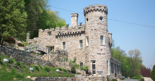 A stone castle with a turret, surrounded by greenery and a pathway, under a clear blue sky.