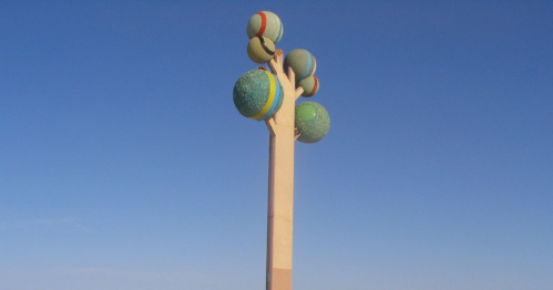 A tall, abstract sculpture resembling a tree with colorful spherical shapes as leaves against a clear blue sky.