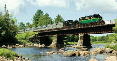 A green and black train crosses a bridge over a river, surrounded by lush greenery and blue skies.