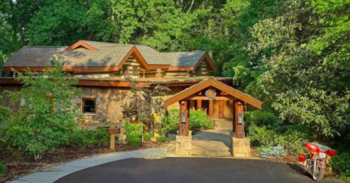A charming stone house surrounded by greenery, featuring a wooden entrance and a red motorcycle parked nearby.