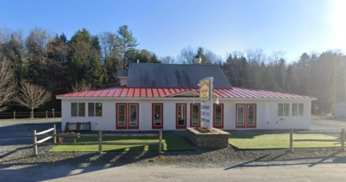A white building with a red roof and trim, surrounded by trees and gravel, featuring a sign in front.