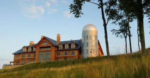 A large rustic lodge with a modern silo, surrounded by tall grass and trees under a clear blue sky.