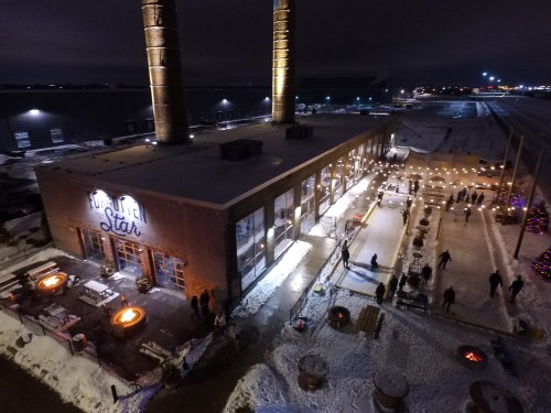 Aerial view of a lively outdoor space with fire pits, snow, and a building with two tall smokestacks at night.