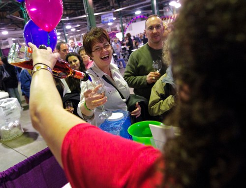 A woman smiles and raises a glass while another person pours a drink at a lively event with balloons in the background.