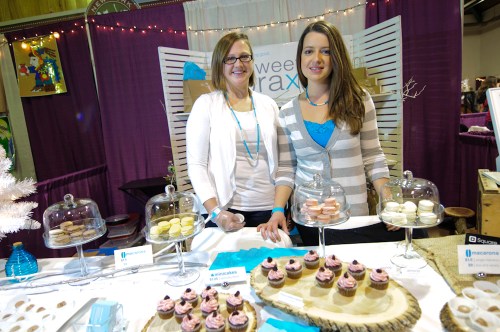 Two women stand behind a dessert table featuring macarons and cupcakes, with a festive backdrop and display cases.