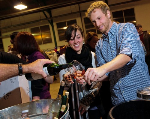 A man and woman smile as they pour drinks from a bottle into a glass at a lively event.
