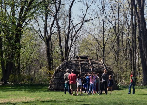 A group of people gathers around a traditional wooden structure in a forested area on a sunny day.