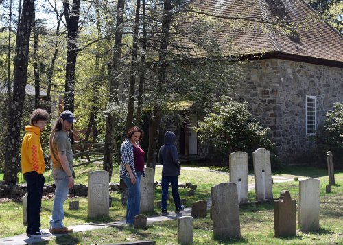 A group of four people stands among gravestones near a stone building, surrounded by trees in a serene setting.