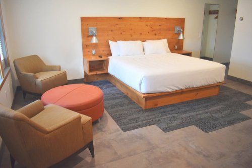 A modern hotel room featuring a queen bed, two chairs, a round ottoman, and wooden accents.